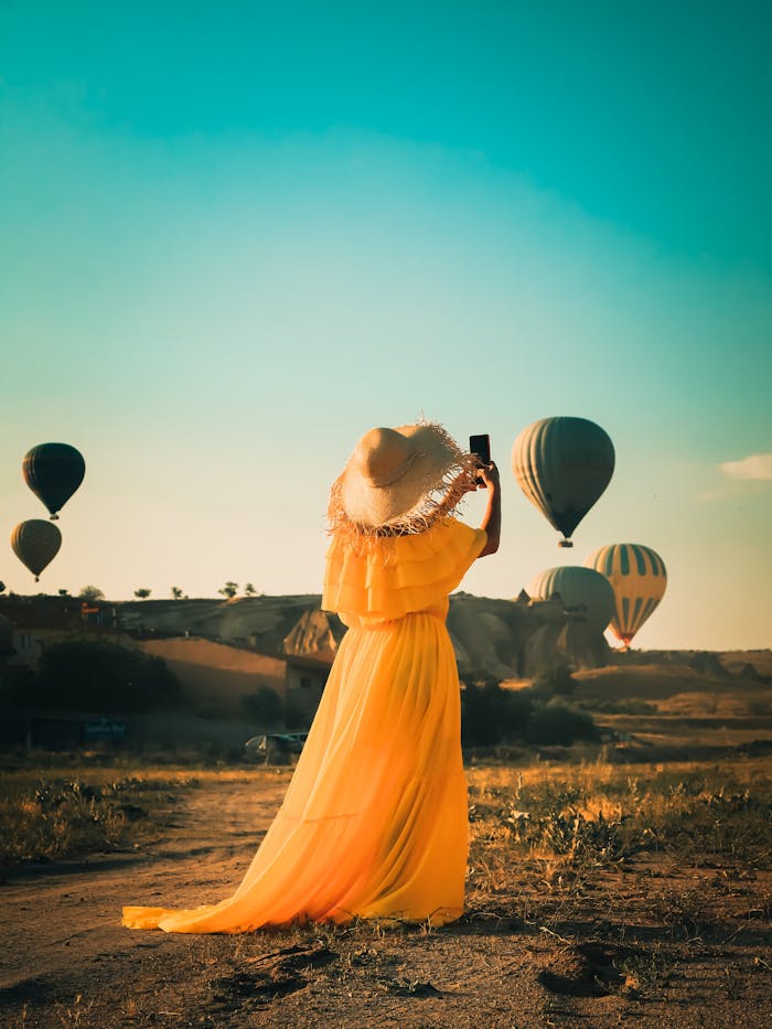 gallery-02 A woman in a yellow dress photographs hot air balloons in a sunny field.