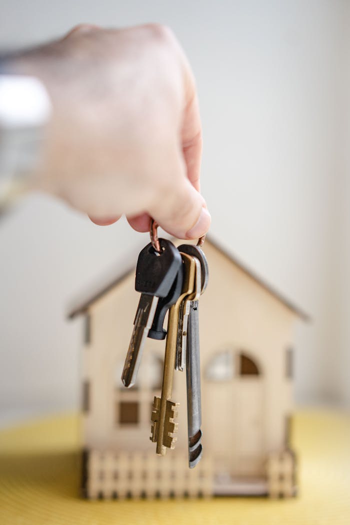 vision Close-up of a hand holding keys with a miniature wooden house in the background, symbolizing real estate investment.