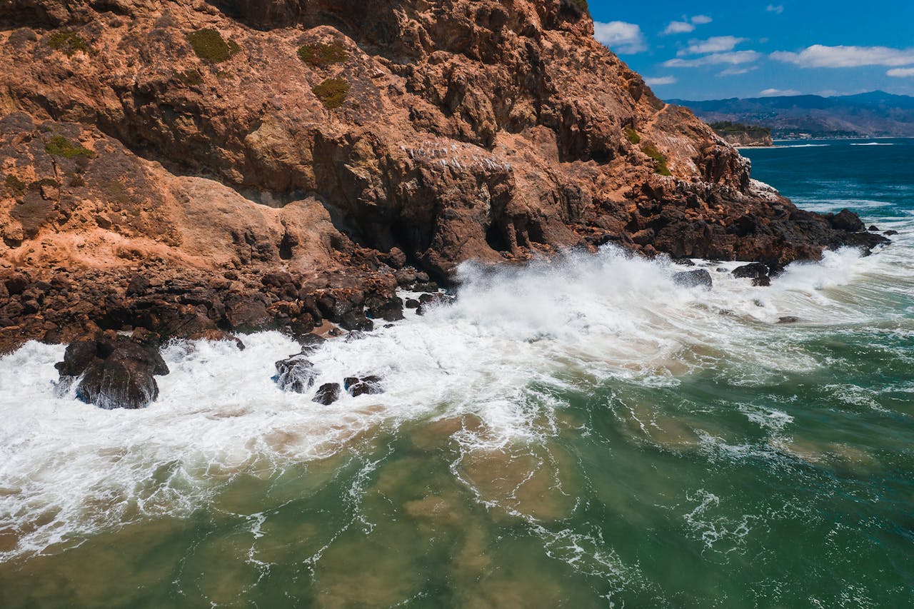 our-story A stunning aerial shot of waves crashing against a rocky shore under a blue sky, capturing the essence of nature's power.